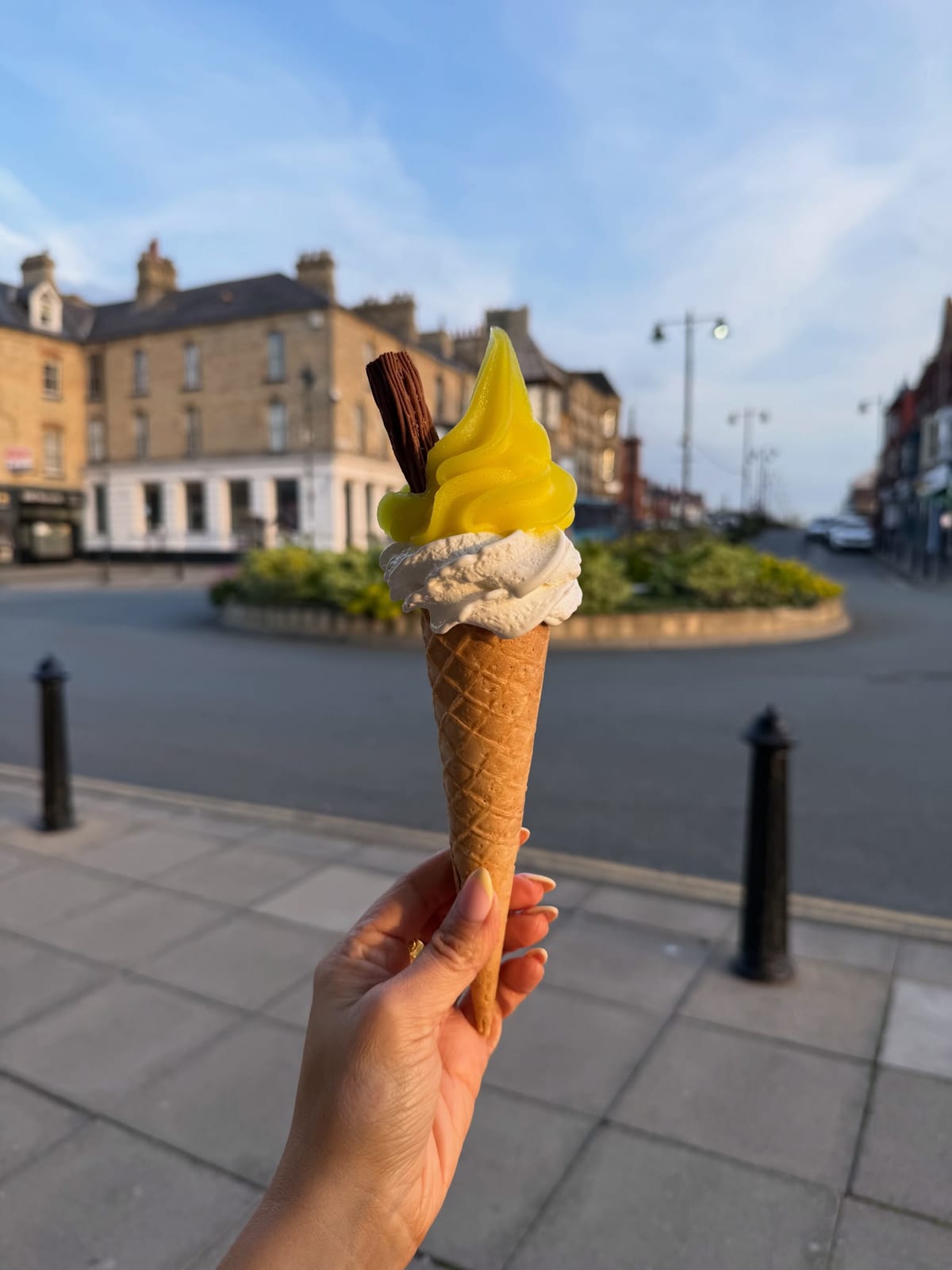 A lemon top whippy ice cream with a Flake, held up in front of Station Square, Saltburn
