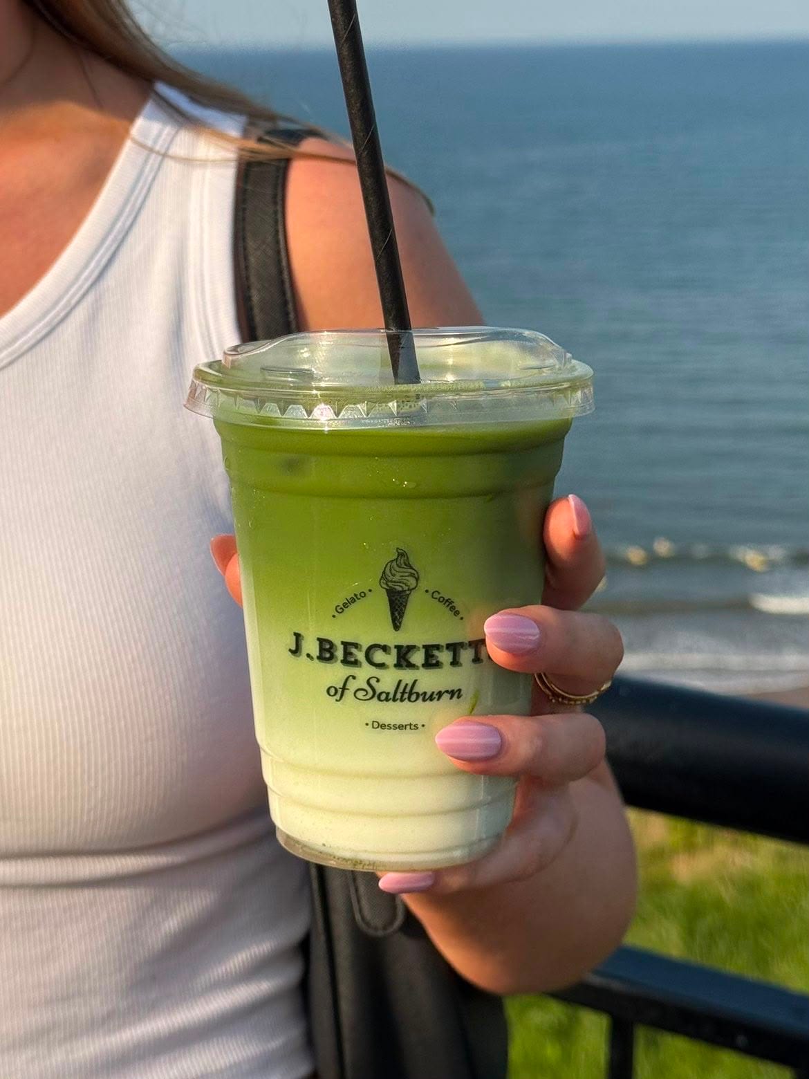 An iced matcha latte in a J.Becketts cup, held up above Saltburn beach