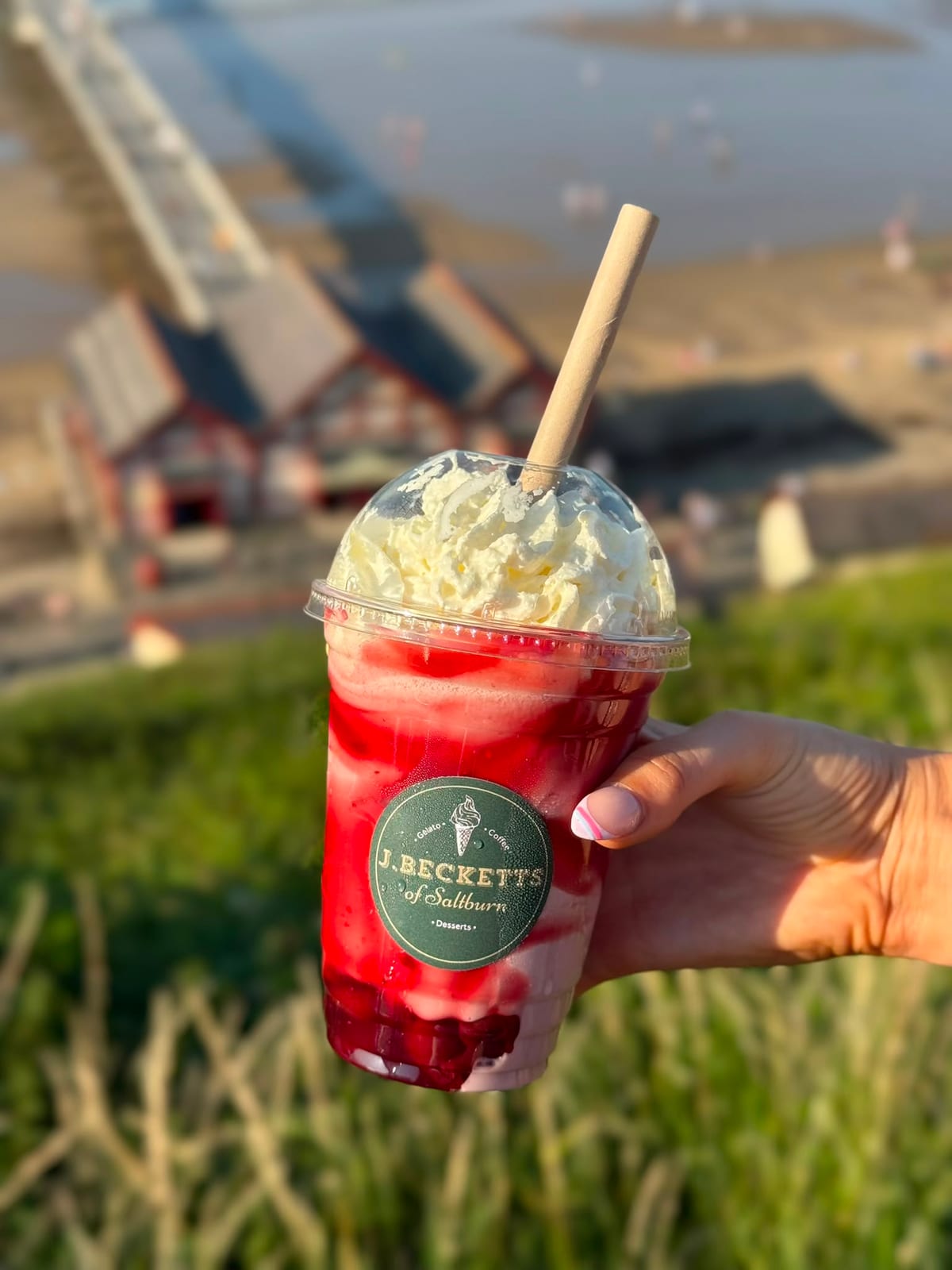 A berry sundae topped with whipped cream, held up above the beach huts and Saltburn pier